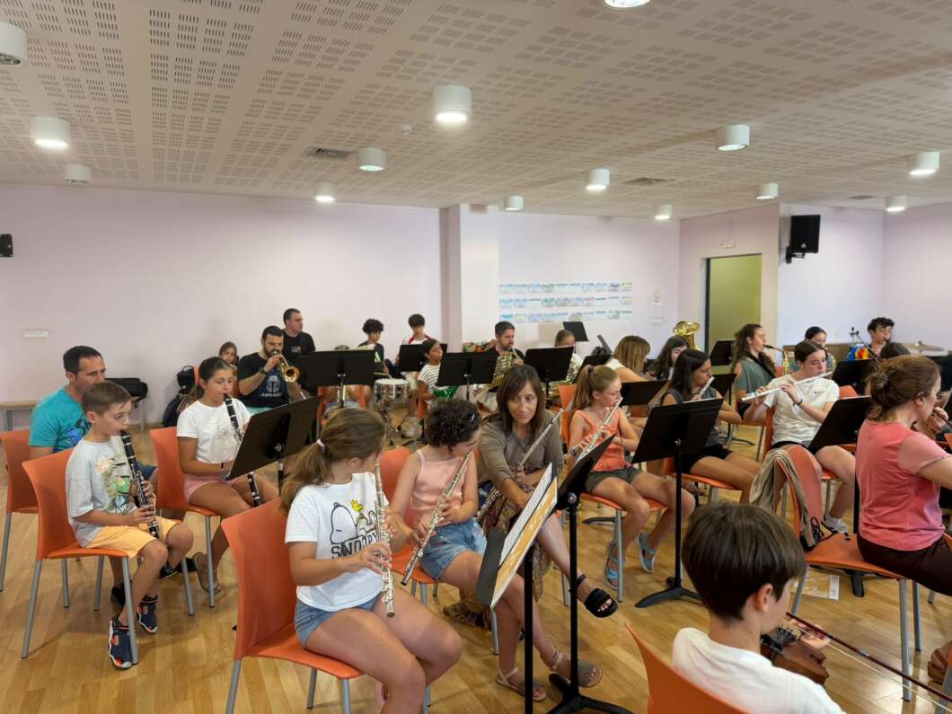 Group of kids in a music room seated on orange chairs, playing wind instruments (flutes, clarinets, brass) at music stands.