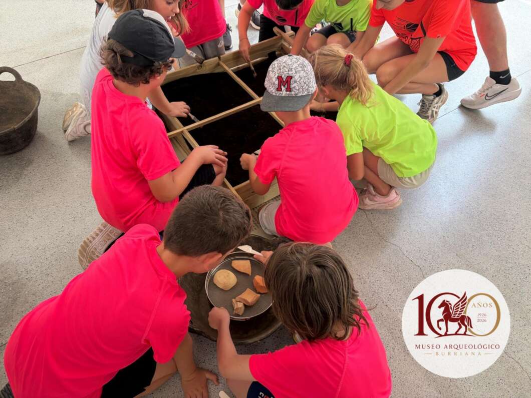 Children in bright pink shirts gather around a dug pit, handling pottery fragments as part of an archaeology activity. Frequently used at a museum education program.