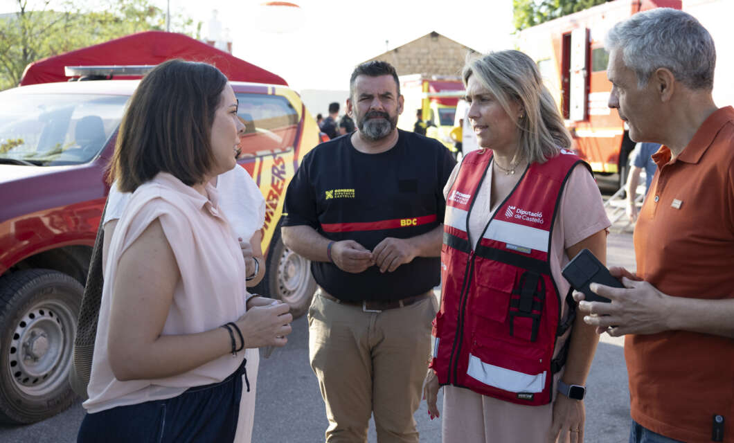 Group of four adults in a disaster response scene, talking near emergency vehicles and a red rescue vest worn by one person.