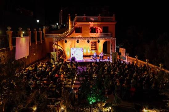 Outdoor nighttime concert on a lit stage built into a villa terrace with an audience seated in rows and warm decorative lighting