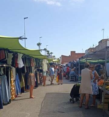 Outdoor market street with green-canopy stalls selling clothes and produce, people strolling on a sunny day.