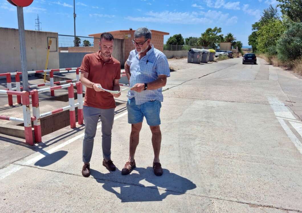 Two men stand on a sunny street, examining a set of papers together near a barrier and curbside.