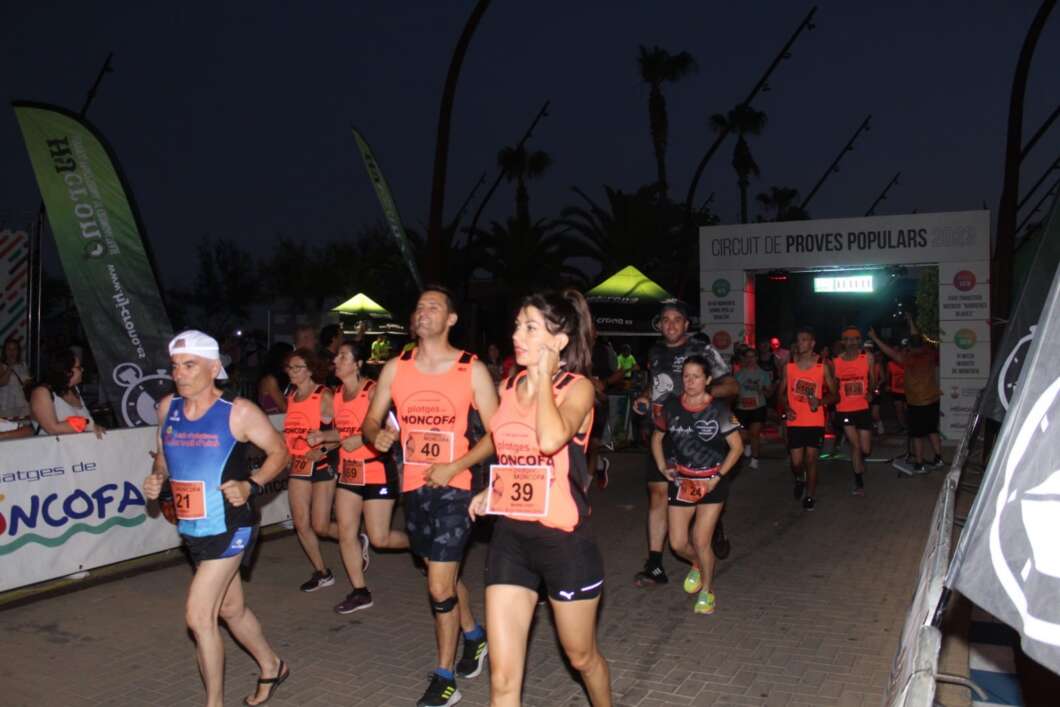 Runners wearing orange bibs sprint through a night-time race, with a finish arch in the background.