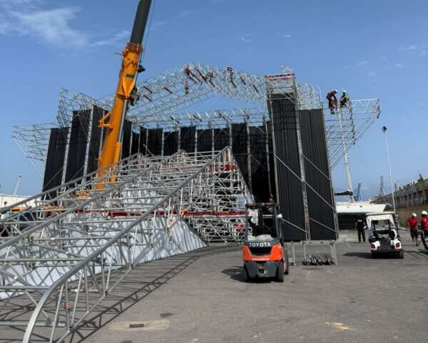 Outdoor construction site with a large metal truss stage under assembly, a crane lifting structure, scaffolding, and workers on the frame.