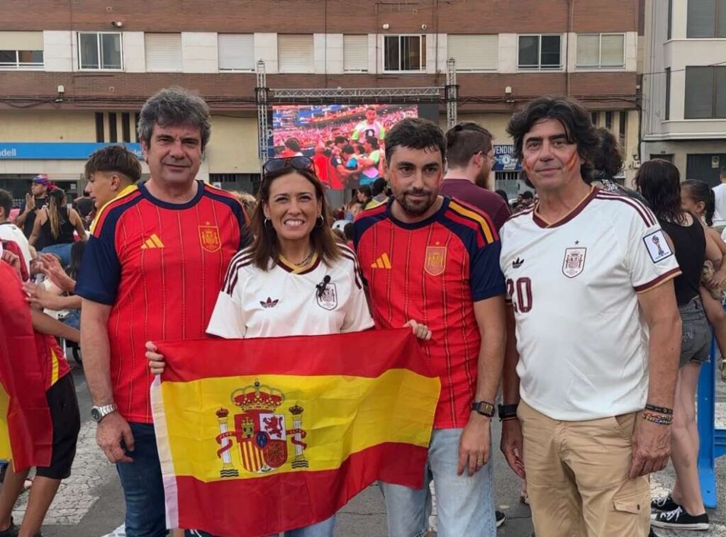 Group of four people wearing Spain soccer jerseys posing together, holding a Spanish flag at an outdoor event with a large screen in the background.