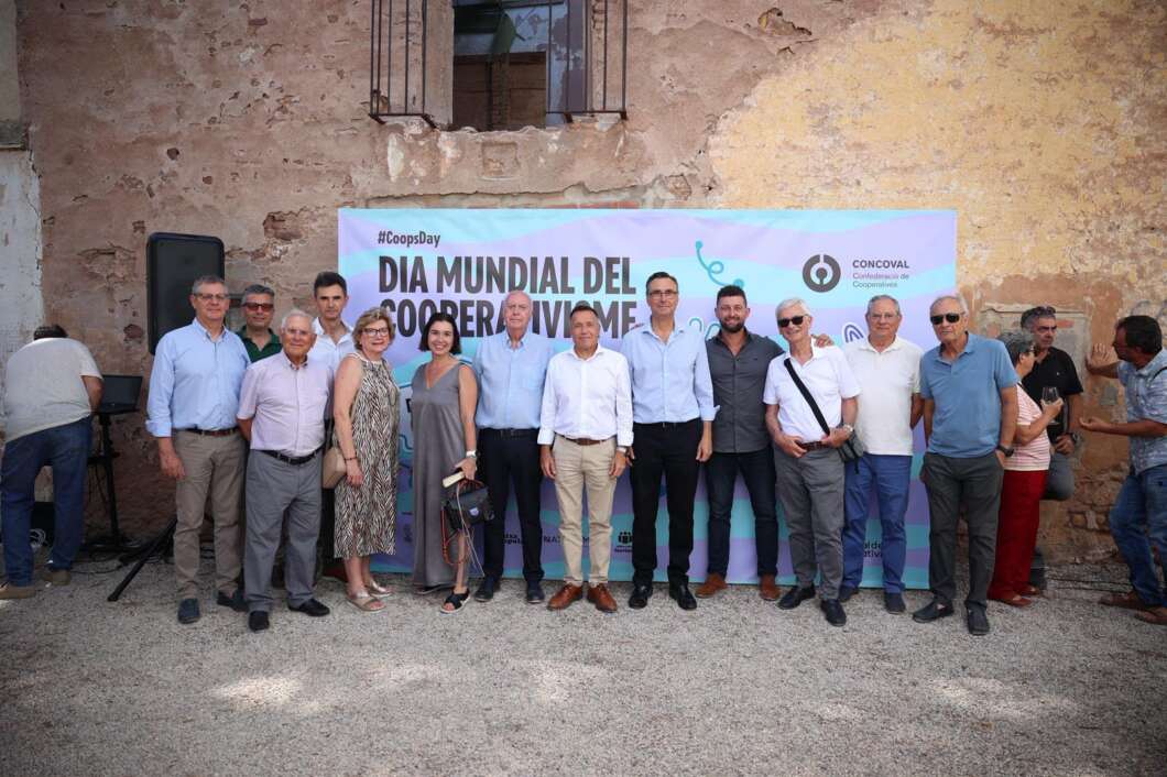 Group of around 15 adults standing side by side outdoors in front of a large event banner, posing for a photo at a formal gathering.