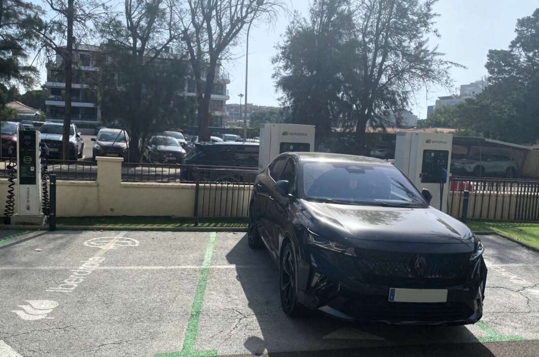 Black SUV parked at an electric vehicle charging station in a lot, with charging kiosks and green-lined parking spaces nearby. The scene shows a sunny day with trees and other cars in the background.