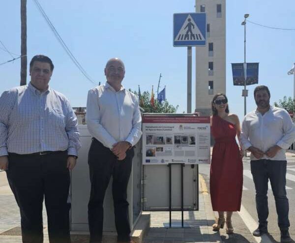 Four adults stand on a sidewalk beside an information board and a pedestrian crossing sign by a tall tower.