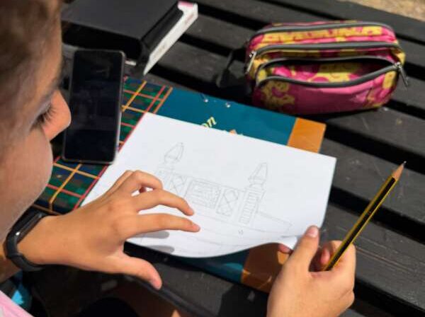 Child rests pencil over a drawing of a rocket on paper at an outdoor picnic table; a pink pencil case, phone, and books nearby.
