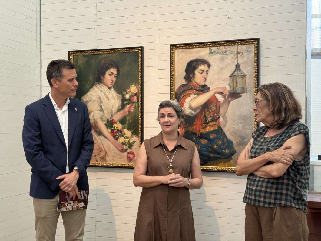 Three adults stand in an art gallery, speaking in front of two large framed paintings of women in period dress.