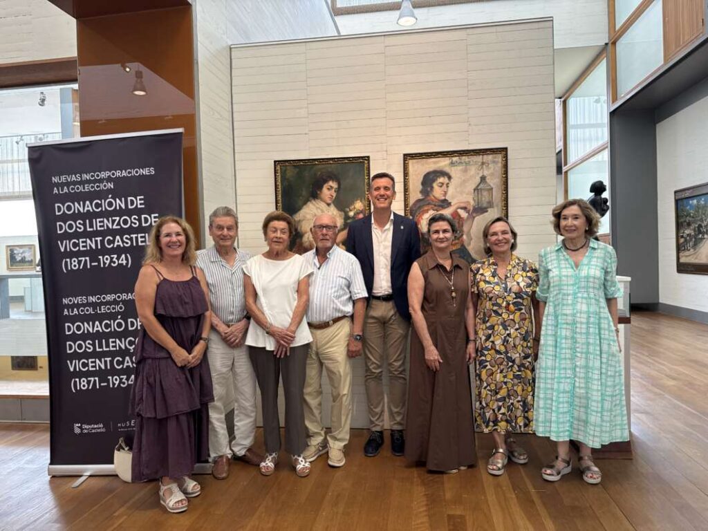 Eight adults pose for a group photo in an art gallery, with two framed paintings behind and a tall exhibition banner on the left.