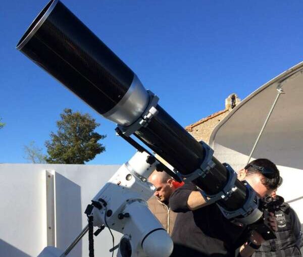 Astronomers adjust a large telescope on an equatorial mount outdoors near a white wall and dome roof.