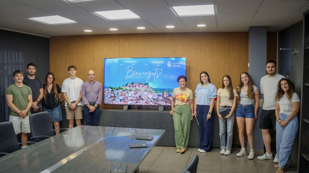 Group of people posing for a welcome photo in a meeting room with a large screen that reads 'Benvinguts!' in the background.