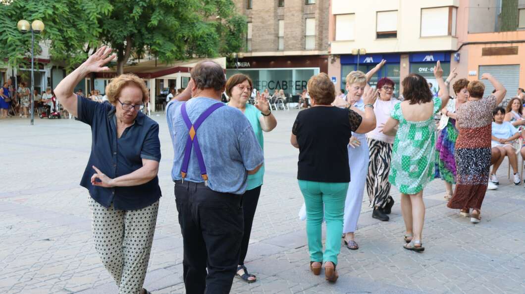 Group of adults dancing together in a sunny town square, some clapping and smiling.