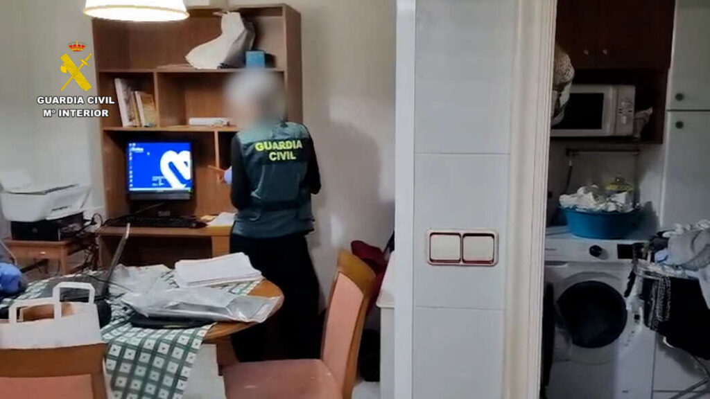 Guardia Civil officer in a dark vest standing at a cluttered desk with a computer in a home office setting.