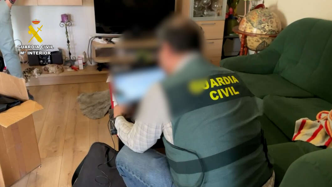 Guardia Civil officer in a green vest helping a seated person at home, logo visible on the left.