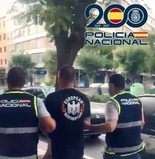 Two Policía Nacional officers escort a man in a black shirt down a city street, with a 200th anniversary logo visible in the upper-right corner.