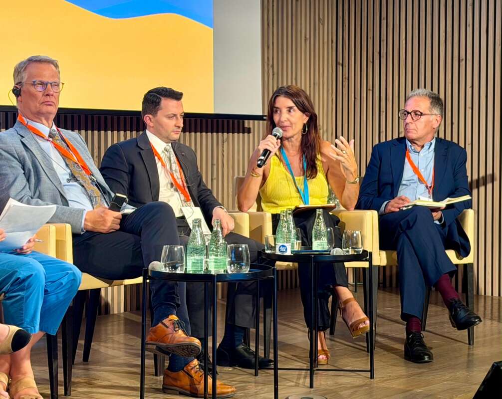 Panel discussion: five professionals seated on stage, woman in yellow speaks into a microphone. (informative)