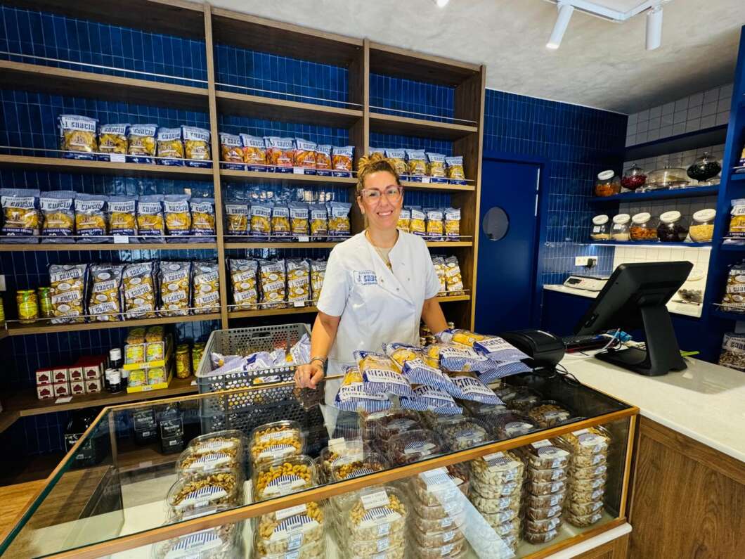 Smiling female clerk in a white uniform behind a glass snack display, with shelves of packaged goods on a blue-tiled wall in the background.