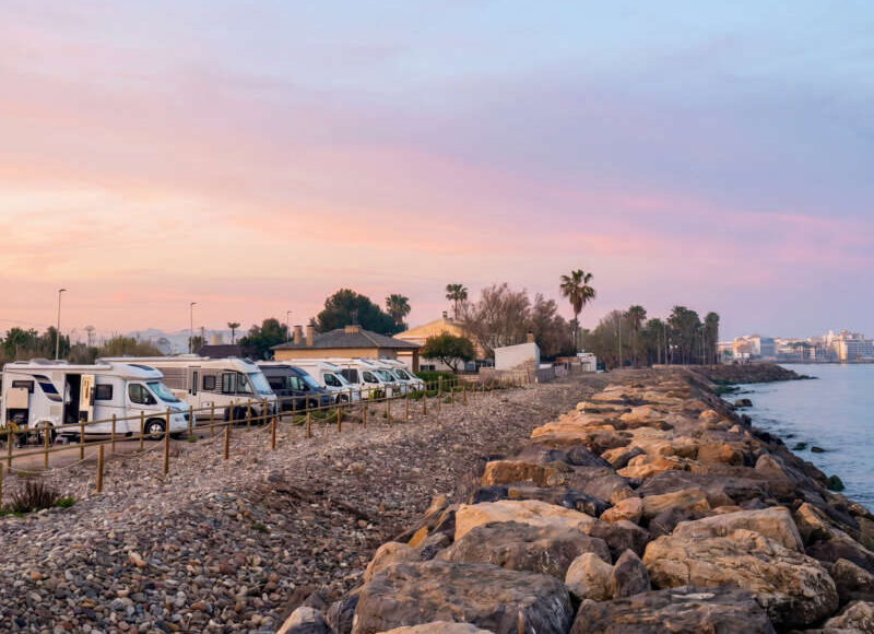 Row of camper vans parked along a rocky shoreline at sunset, with a pastel pink–blue sky and calm water to the right.