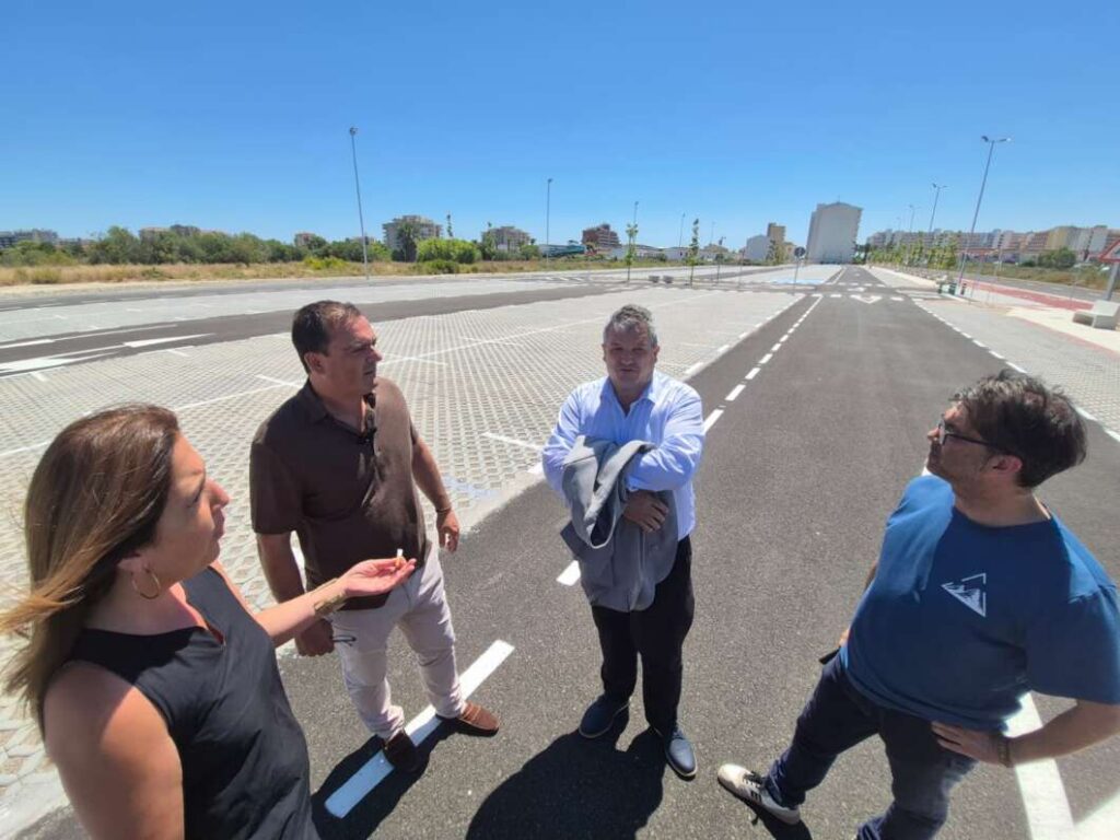 Four adults stand in the middle of a wide, freshly paved road on a sunny day, talking together.