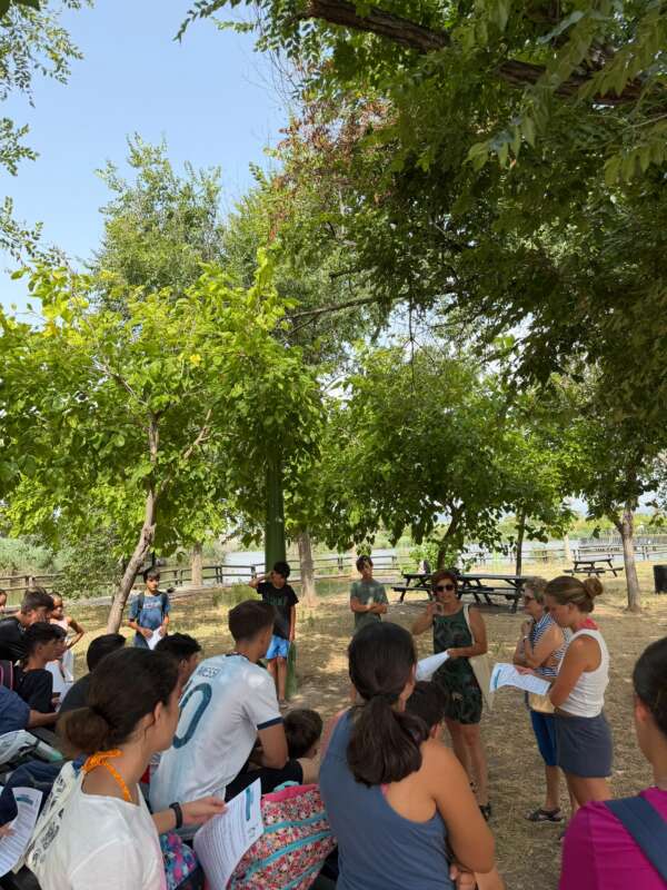Group of children and adults gathered in a park under trees, standing in a circle and reading from papers as a facilitator leads the activity.