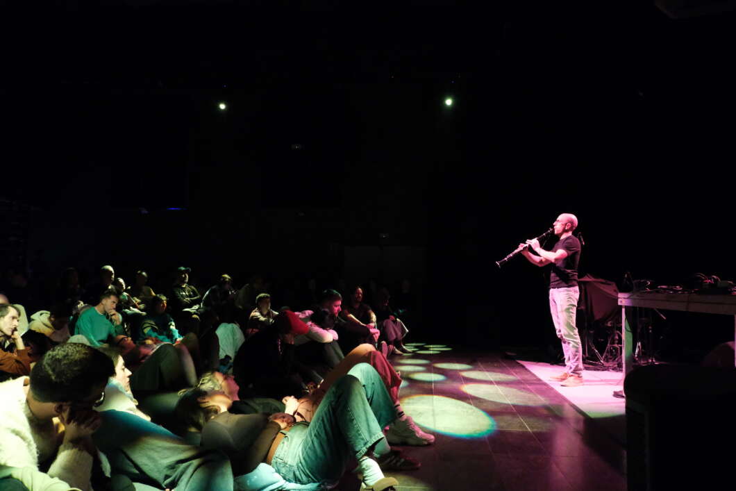 A musician plays a clarinet on stage while a dimly lit audience sits on the floor watching.