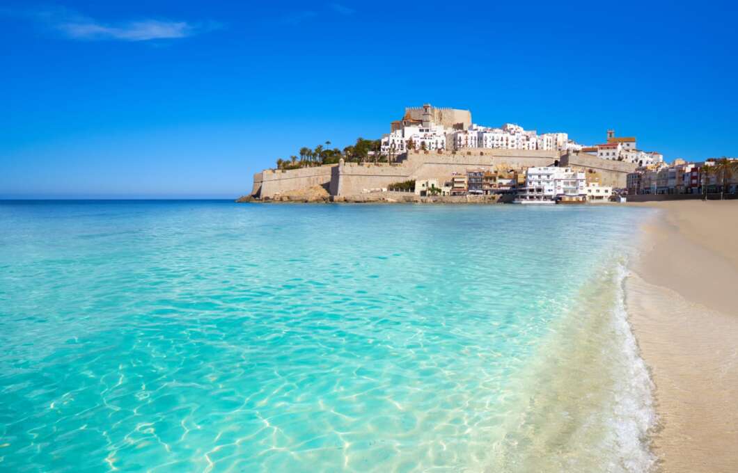 Turquoise sea meets a sandy beach with a historic fortress and white buildings atop a rocky promontory under a clear blue sky.