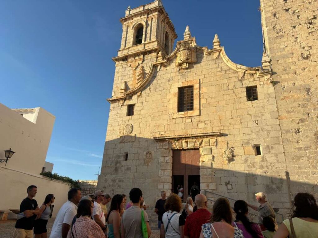 Stone church facade with a decorative doorway and bell tower, as a group of tourists gathers outside on a sunny day outside the historic building.