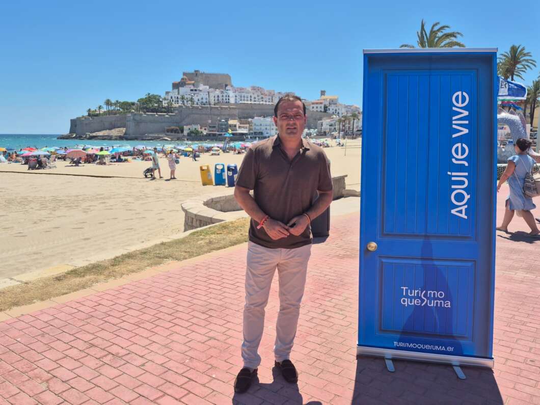 Man stands on a sunny beach next to a tall blue door with the words 'Aqui se vive' and a coastal town in the background.