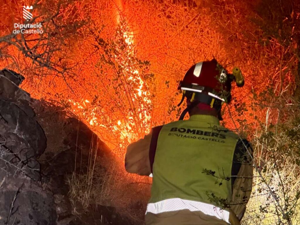 Firefighter in a protective vest and helmet facing a blazing brush fire at night, with the Diputació de Castelló logo visible in the corner.