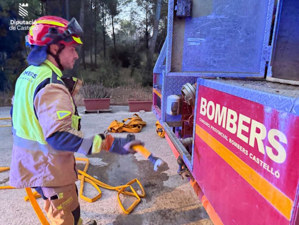 Firefighter in a yellow-vested suit and red helmet beside a blue fire truck, handing a strap with hoses on the ground.