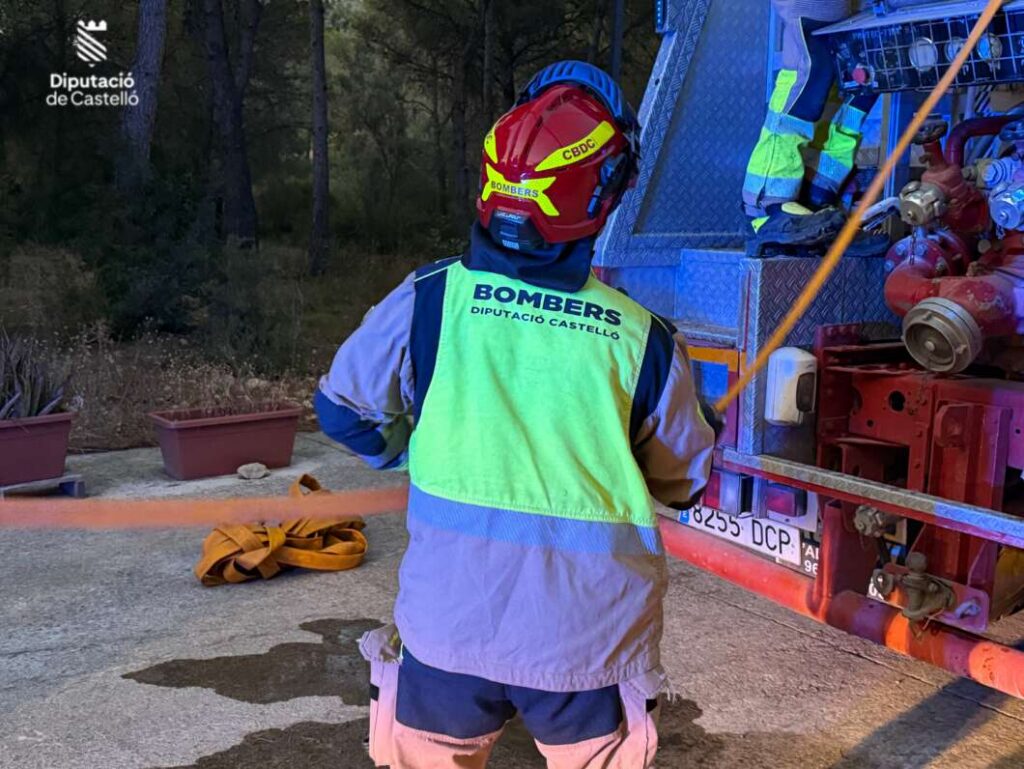 Firefighter in a bright high-visibility vest and red helmet labeled BOMBERS, standing beside a fire engine in a forested area.