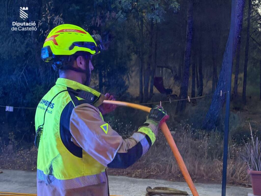 Firefighter in a neon yellow helmet and jacket handling an orange hose outdoors, with a forested area in the background and a Castellón government logo in the corner.