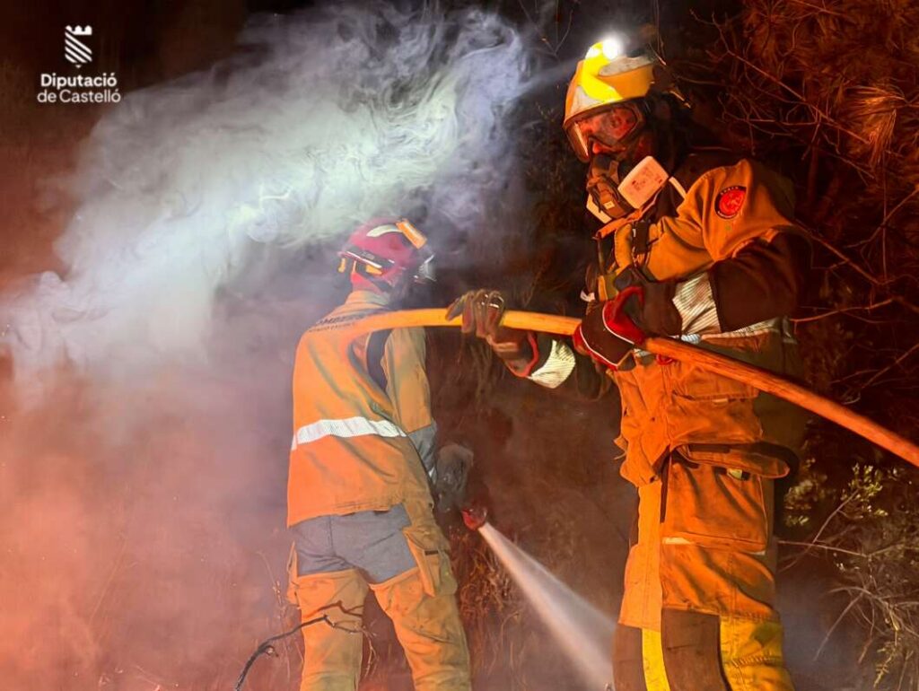 Two firefighters in full turnout gear spray water on a night fire, smoke rising around them, logo in the corner.
