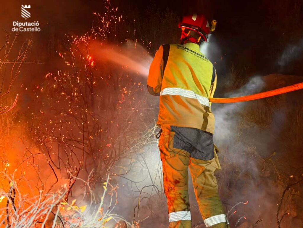 Back view of a firefighter in high-visibility gear spraying water on a brush fire at night, sparks and smoke around