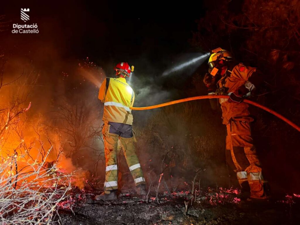 Night scene of two firefighters in yellow turnout gear battling a forest fire, directing a hose amid flames and smoke, with a Diputació de Castelló logo in the corner.