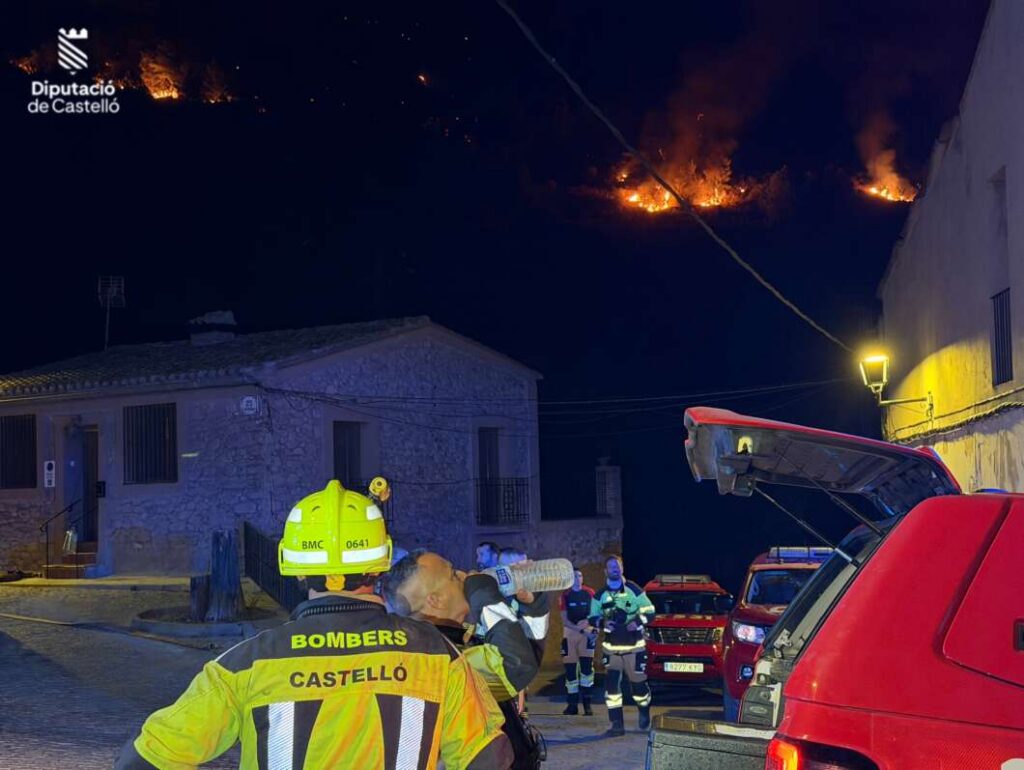 Firefighters in high-visibility gear at night, battling hillside flames above a stone house in Castellón with emergency vehicles nearby