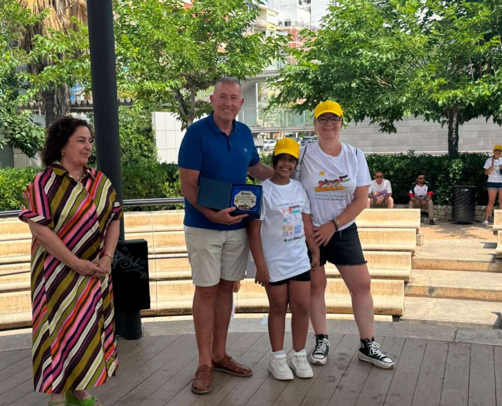 Group of four people posing outdoors; man in blue shirt holds an award plaque beside a young girl and woman wearing yellow caps and matching shirts, with an older woman in a striped dress watching on the left.