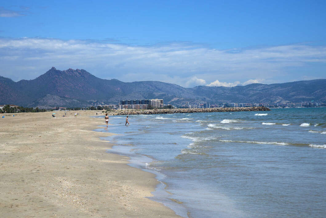 Beach scene with two people walking along the water's edge, calm waves, and distant mountains and buildings under a blue sky.