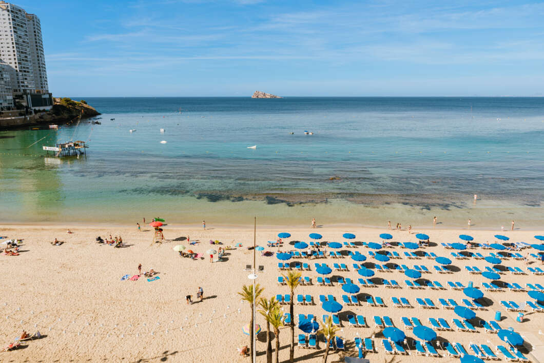 Busy sandy beach with rows of blue umbrellas and chairs, calm turquoise water, and tall buildings on the left; a distant rocky island sits on the horizon.