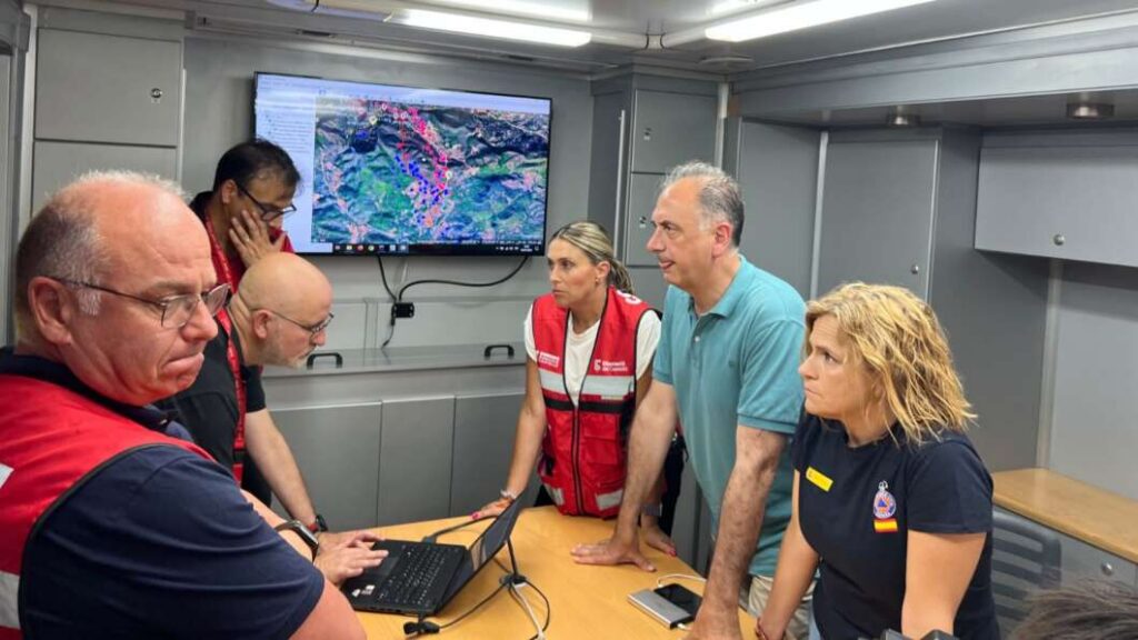 Team of responders in red vests gather around a laptop inside a mobile command center, reviewing a map displayed on a wall monitor.
