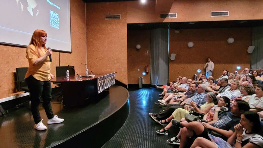 Woman in an orange shirt speaks into a microphone on a stage before a seated audience in a lecture hall with a projection screen to the left.