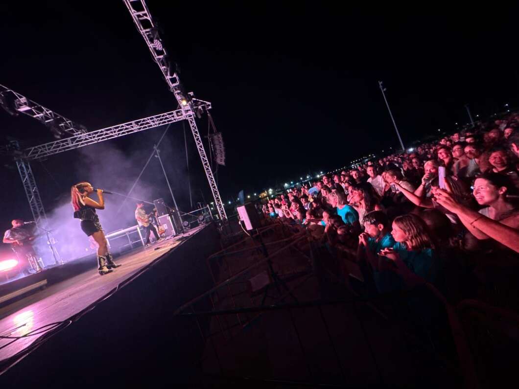 Female singer on stage at night performing to a cheering crowd, with stage lights and scaffolding overhead.