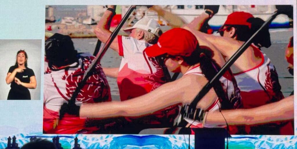 Dragon boat team in red and white uniforms paddling in unison on the water, with a sign-language interpreter shown in a small inset on the left.