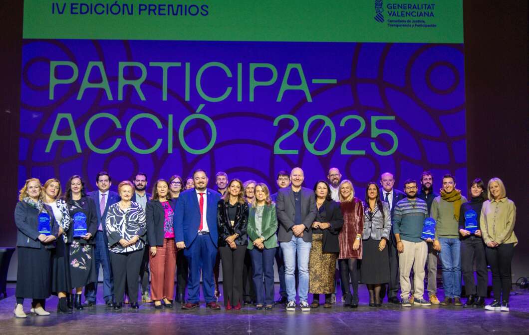 Grupo de unas 25 personas en un escenario en la IV edición de Premios Participa-Acció 2025, algunos con trofeos azules, fondo morado.