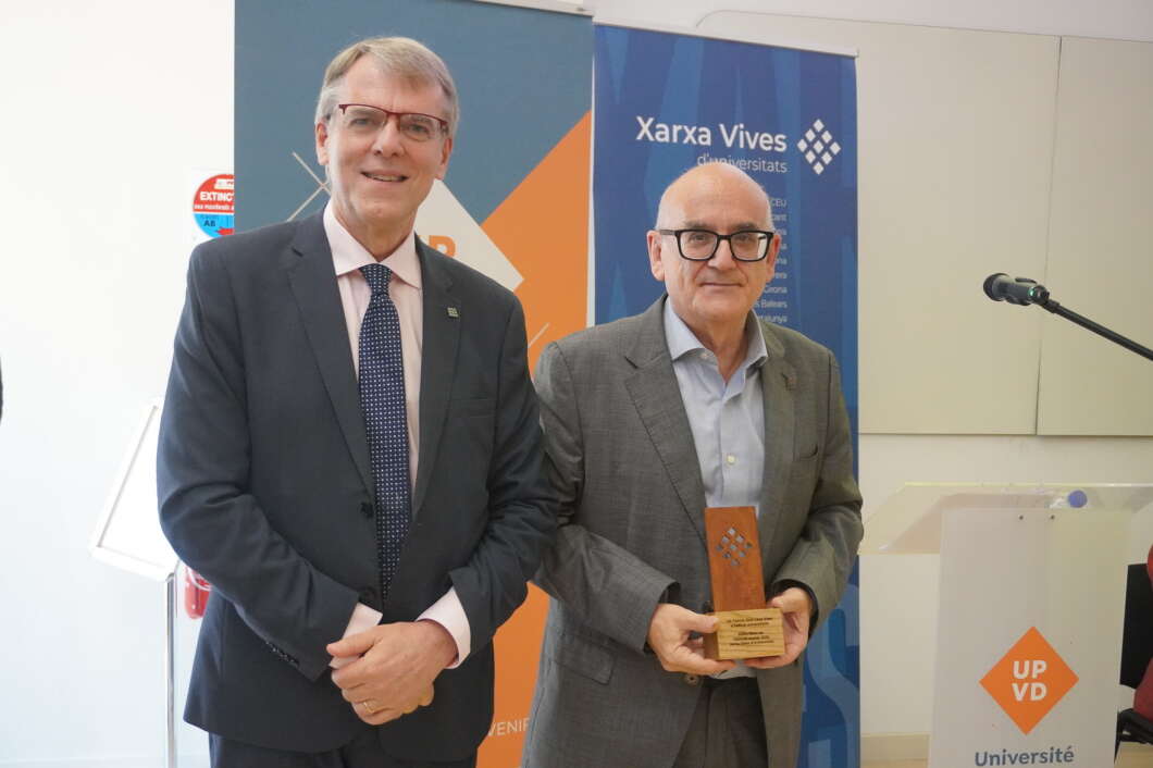 Two suited men at an award ceremony; the man on the right holds a wooden trophy, with banners behind them.