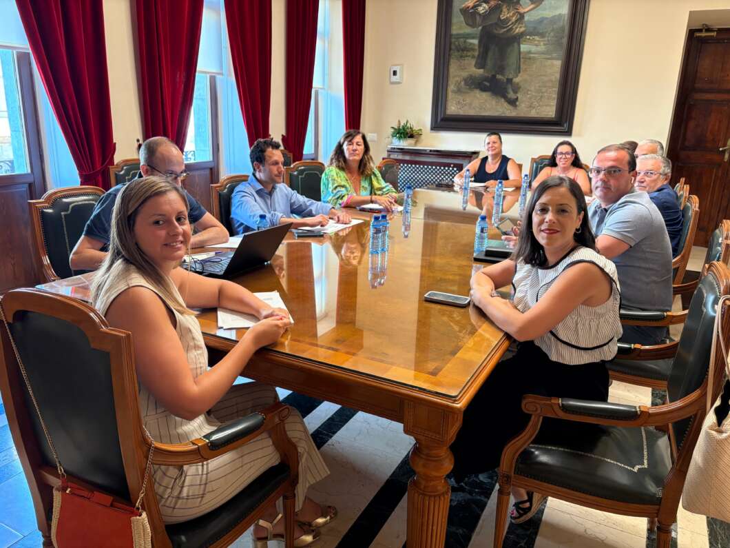 Group of professionals seated around a large wooden conference table in a sunlit room with red drapes and a framed painting on the wall.