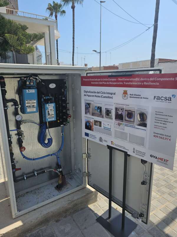 Outdoor cabinet housing water-treatment equipment with blue hoses and gauges, placed beside a large project information board about the digitalization of the water cycle in Nules, Castellón, Spain.