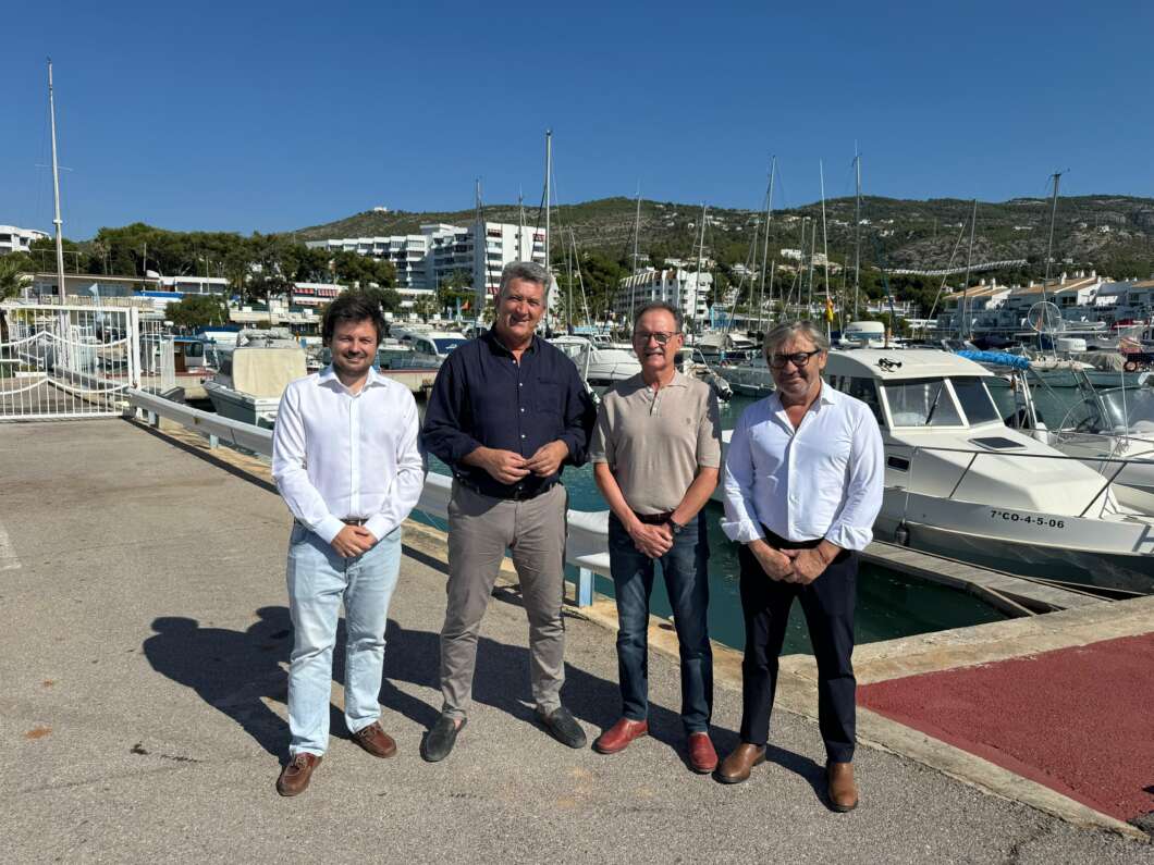 Four men stand side by side on a marina promenade, with sailboats and a harbor behind them on a sunny day.